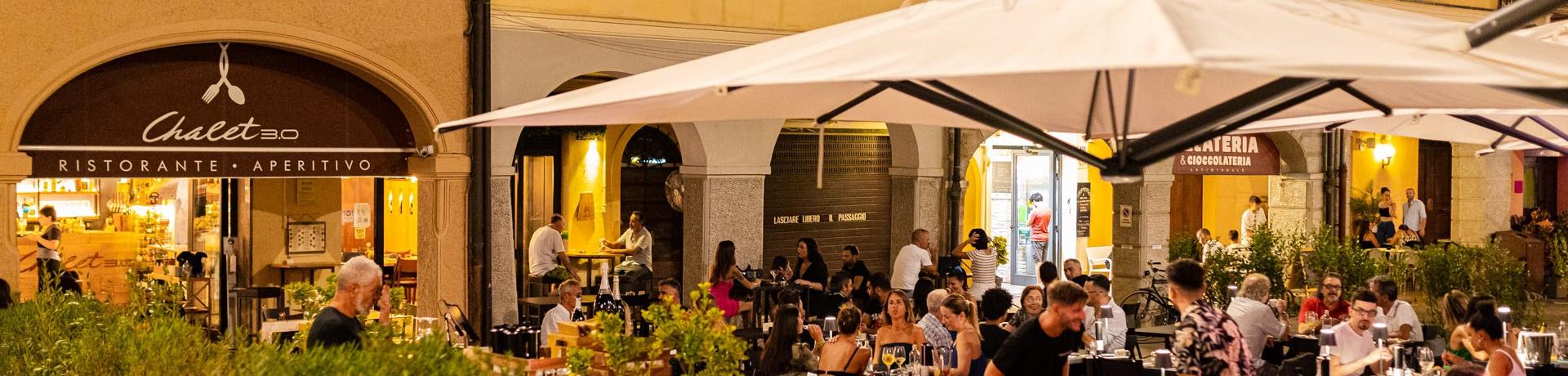 Ristorante all'aperto affollato di sera, sotto grandi ombrelloni, atmosfera vivace e accogliente.