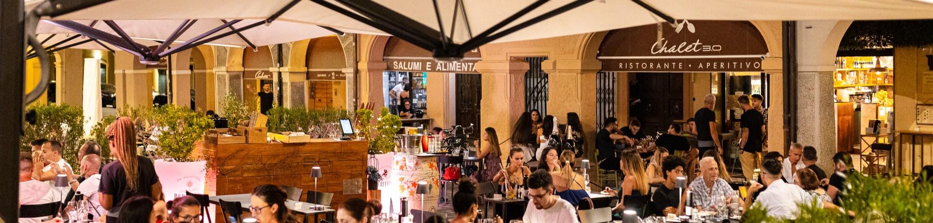 Ristorante all'aperto affollato di sera, con persone che cenano e chiacchierano.