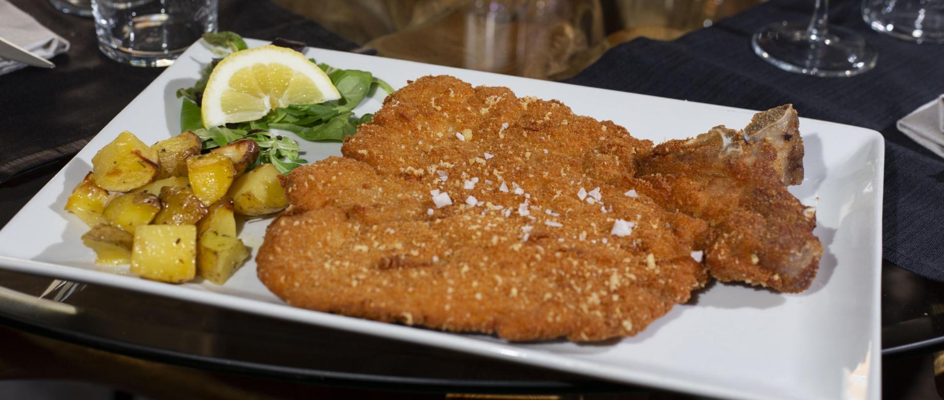 Cotoletta impanata con patate arrosto, insalata e limone su un piatto bianco.